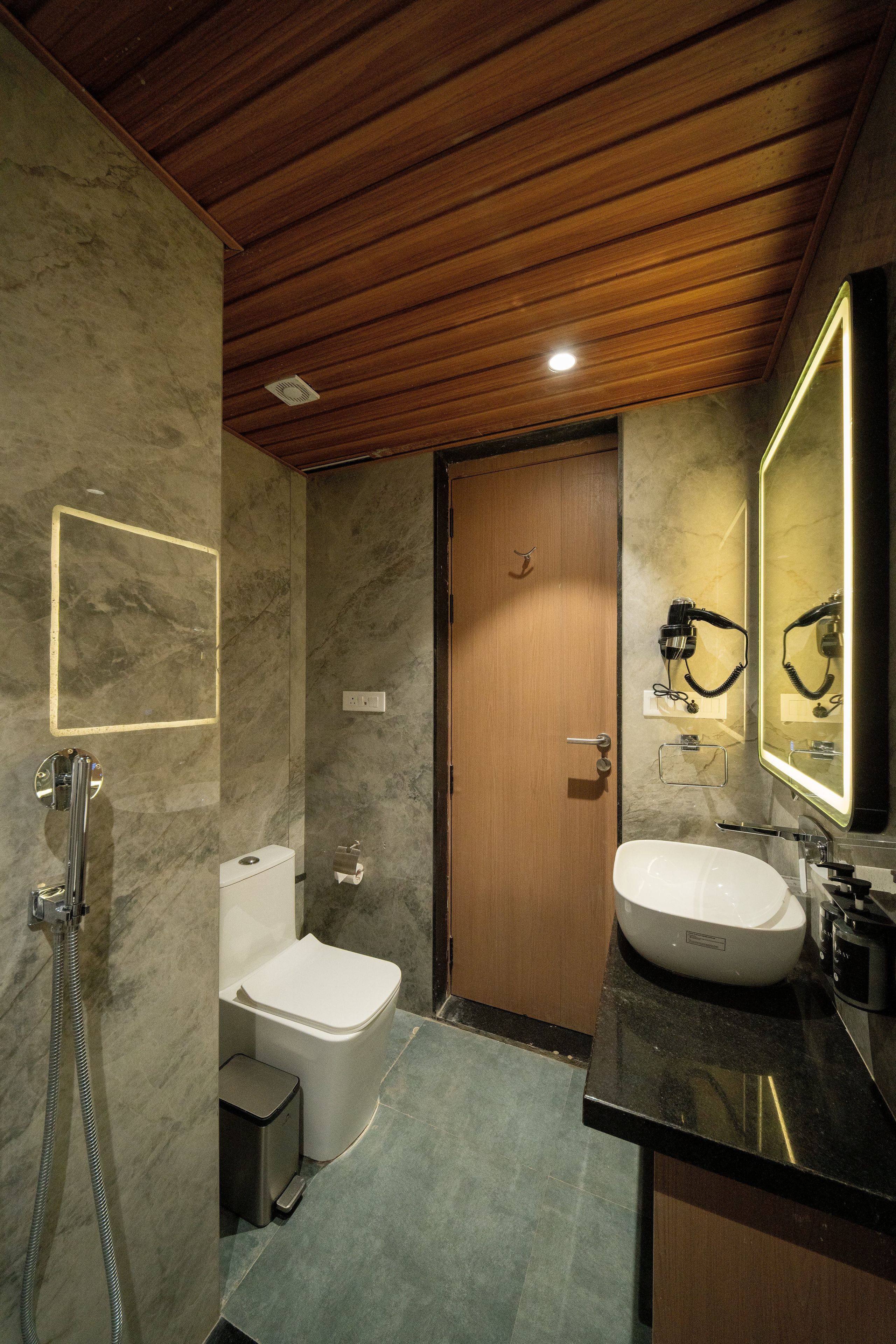 A modern bathroom with a wooden ceiling, a marble wall, a wooden door, a white toilet, a sink with a mirror, and a shower.