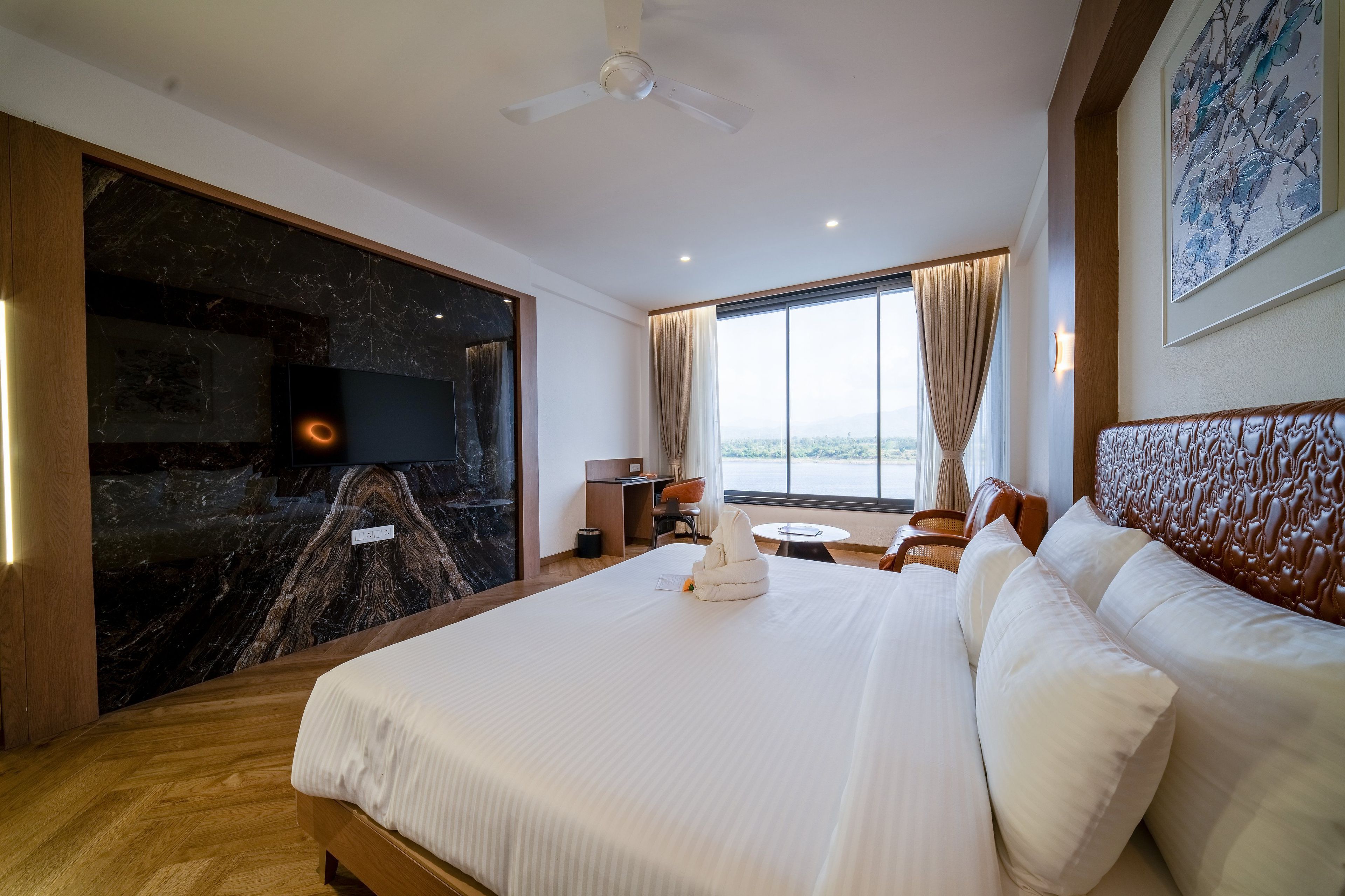 A hotel room with a large bed, a flat-screen TV mounted on a marble wall, a desk with a chair, and a view of the outdoors through a large window.
