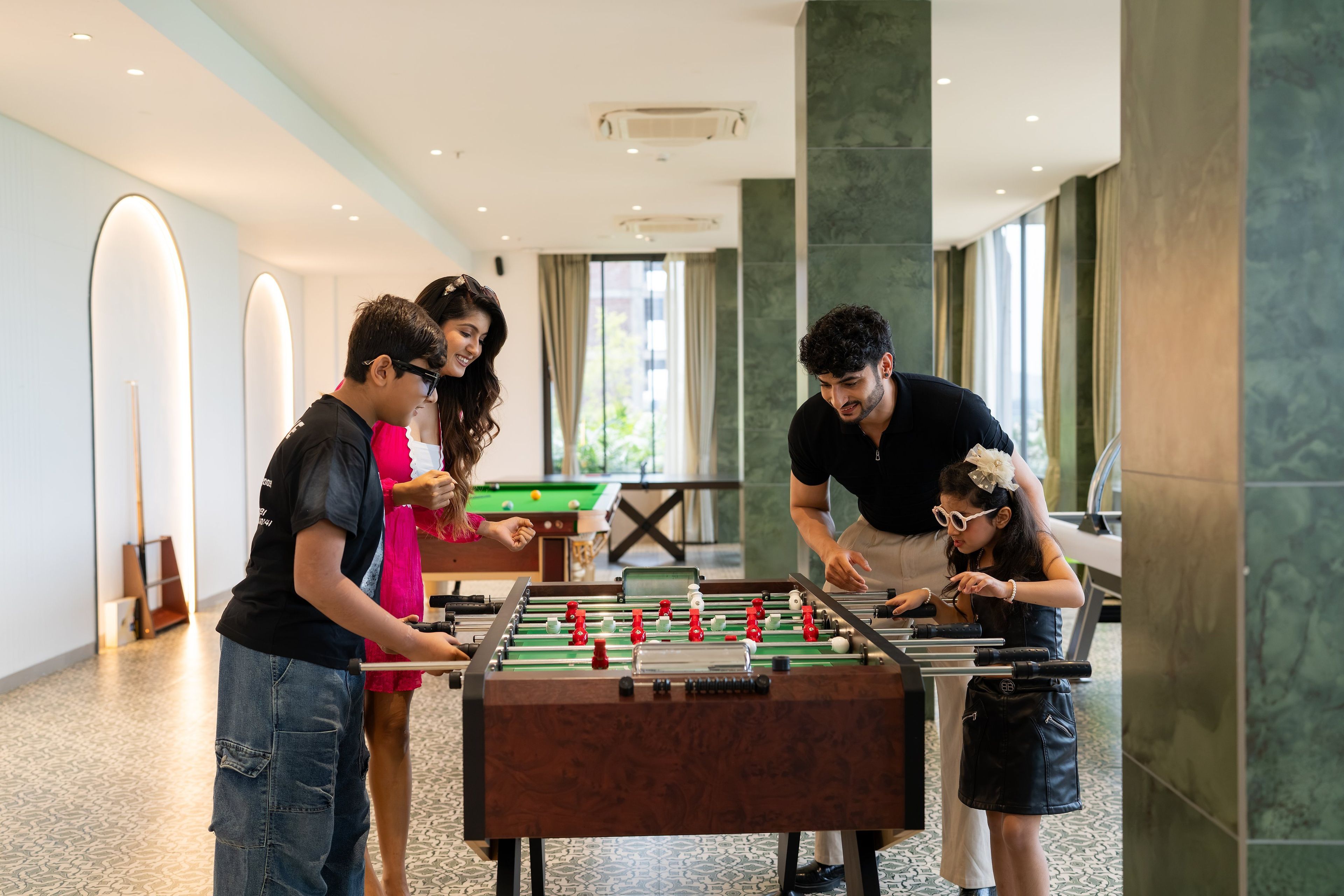 A group of people playing foosball in a well-lit indoor space with a pool table in the background.
