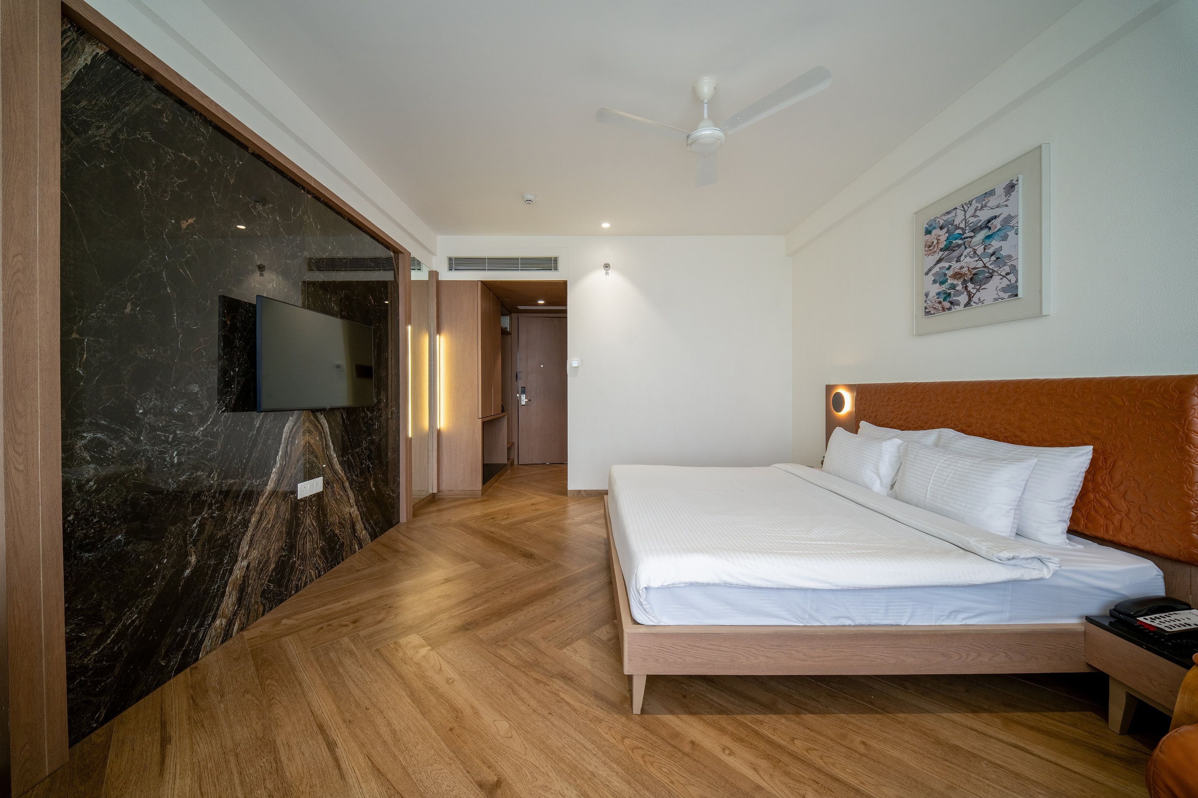 A hotel room with a bed, a television, a ceiling fan, and a painting on the wall.
