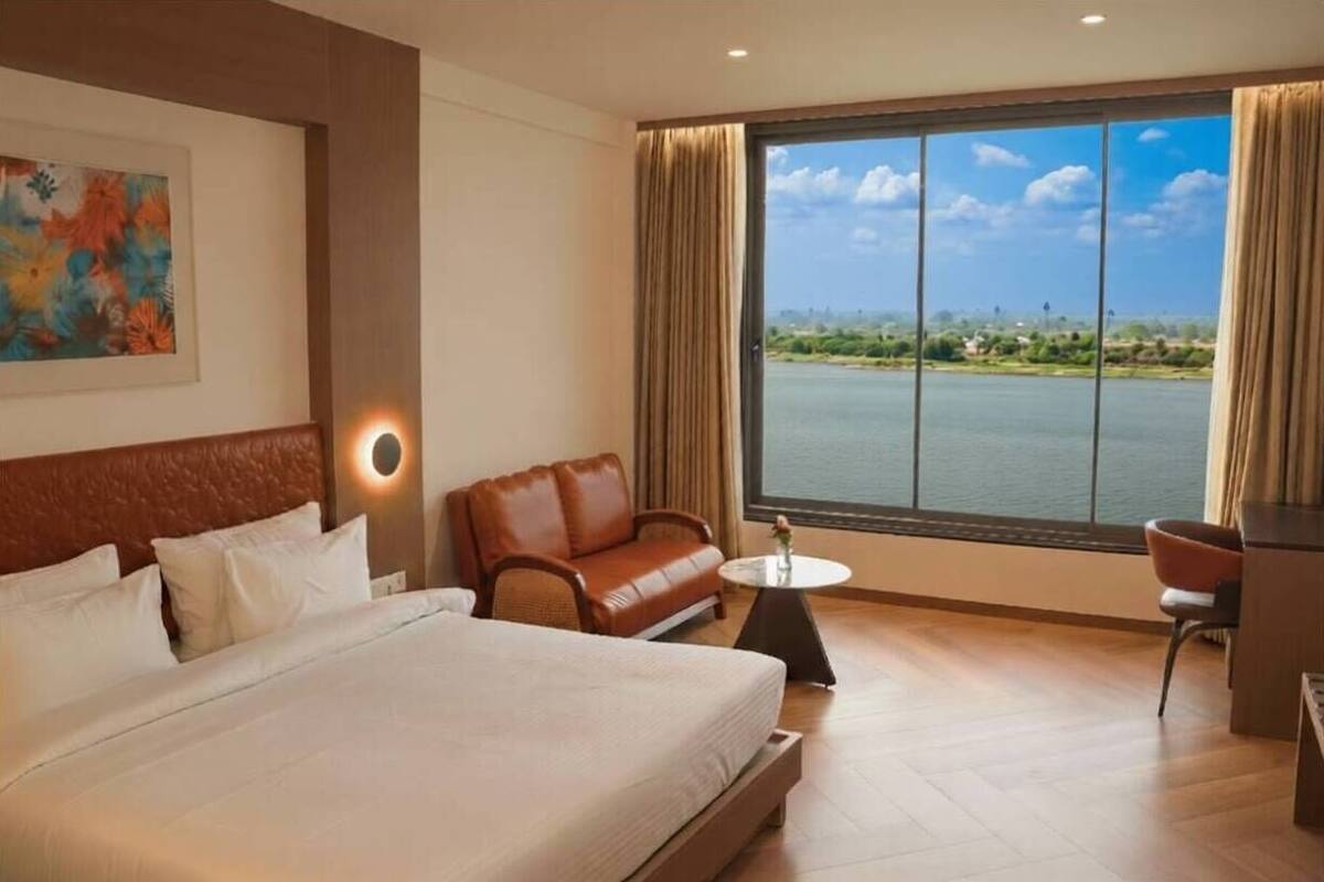 Une chambre d’hôtel avec un grand lit, un canapé en cuir marron, une petite table ronde et une vue sur une étendue d’eau, visible à travers une grande fenêtre.