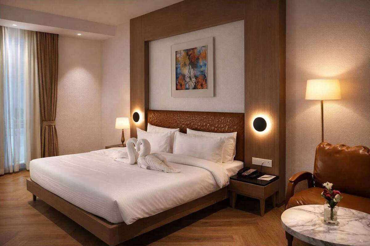Une chambre d’hôtel avec un grand lit, deux lampes de chevet, une tête de lit dotée d’un éclairage circulaire, un tableau au-dessus du lit, un fauteuil en cuir marron et une petite table avec un vase.