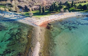 Eine Luftaufnahme einer Küstenregion mit klarem Wasser, einem Sandstrand und einem Feldweg, der zu einer grünen, von Bäumen gesäumten Fläche führt.