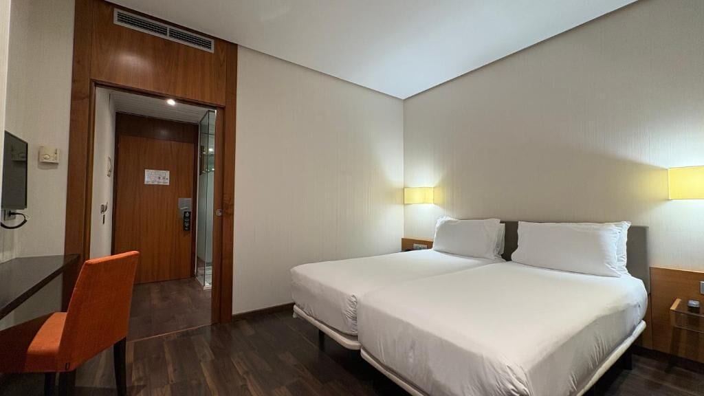 Habitación de hotel con una cama grande, un escritorio y una silla. Se ve una puerta que da al baño y un televisor montado en la pared.