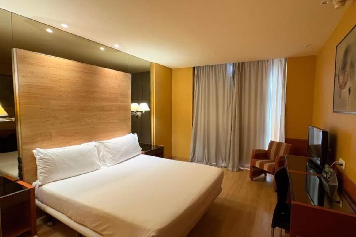 Een hotelkamer met een groot bed, een bureau met stoel, een televisie en een groot raam met gordijnen.