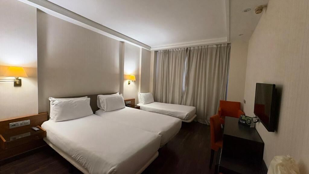 A hotel room with two beds, a desk, a chair, a TV, and a window with curtains.