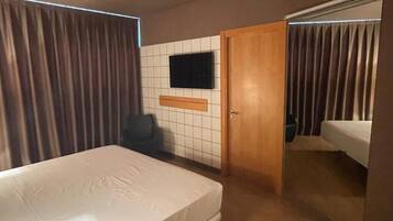 Habitación de hotel con una cama individual, televisor montado en la pared, una puerta que da a otra habitación y una ventana con cortinas.