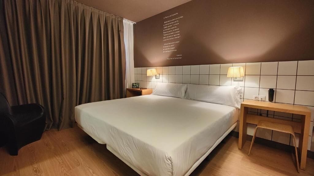Hotellihuone, jossa on suuri sänky, kaksi yöpöytävalaisinta, puinen yöpöytä, pieni pöytä tuolilla sekä musta tuoli.