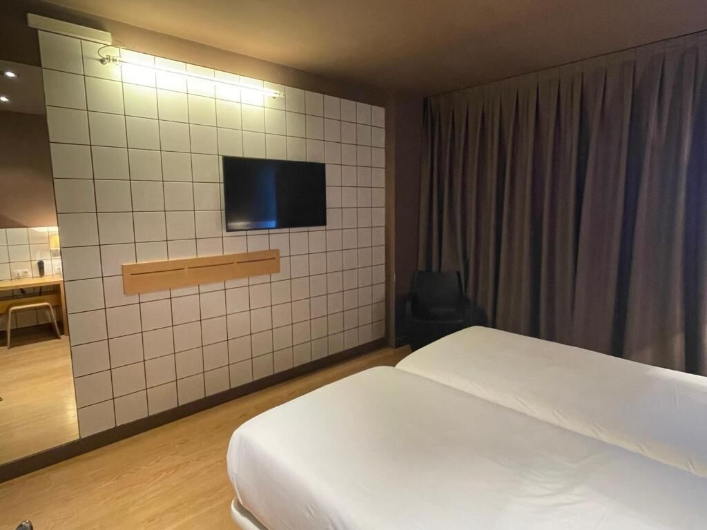 A hotel room with a large bed, a flat-screen TV mounted on a tiled wall, a desk with a chair, and a window with curtains.