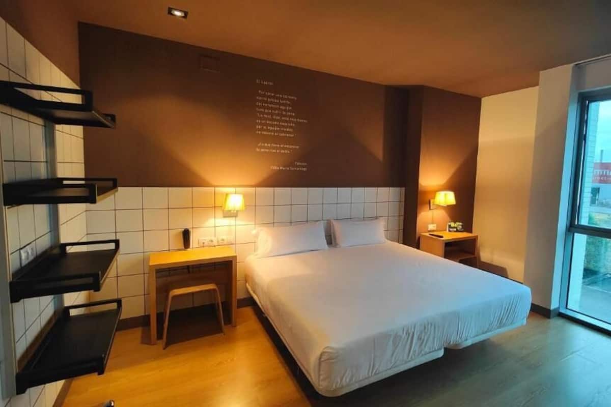 A modern hotel room with a large bed, bedside tables, a small stool, and a view of the outside through a large window.