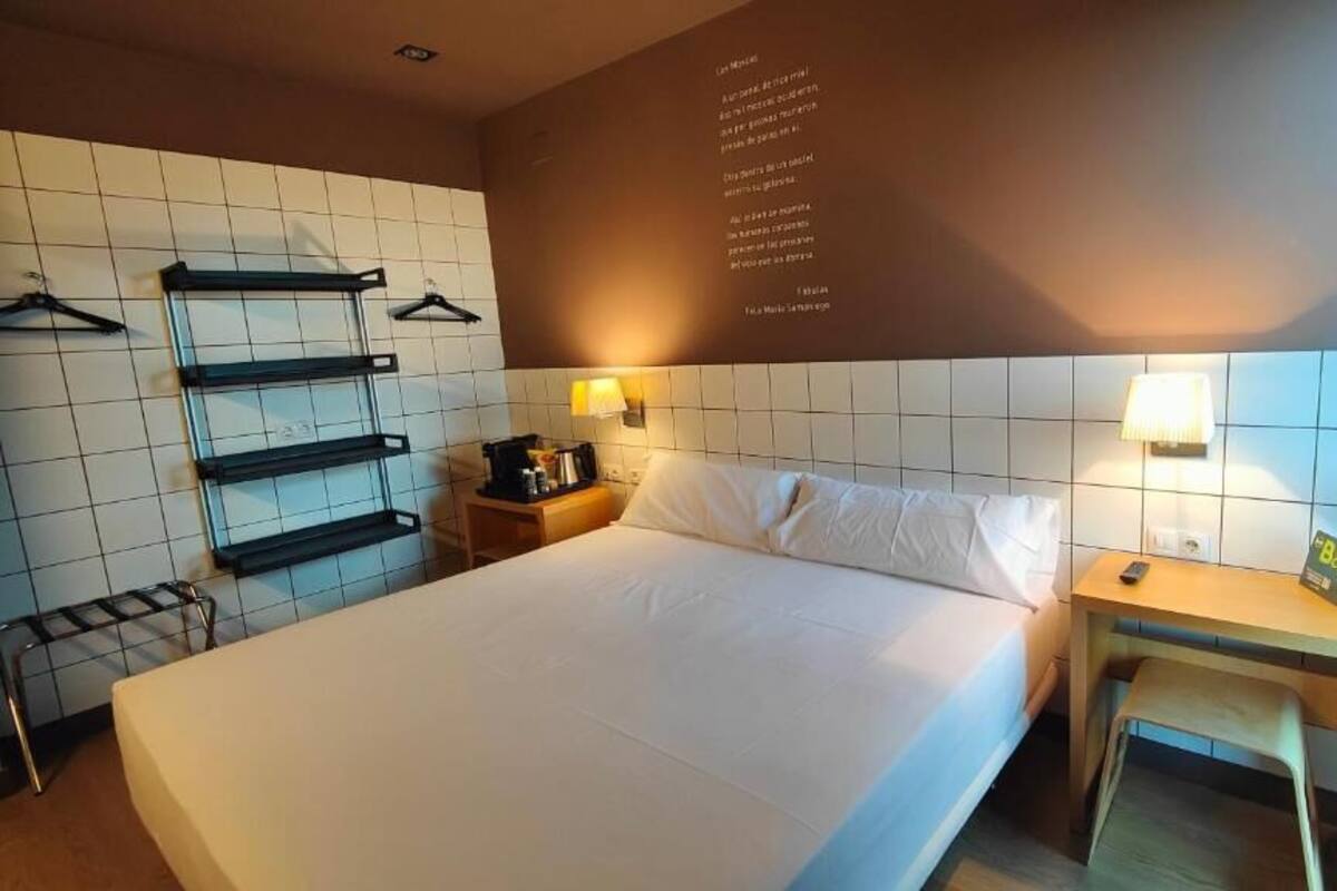 A hotel room with a bed, a desk, a chair, a nightstand, a lamp, and a wall-mounted shelf with hangers.