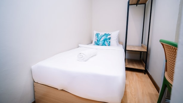 Una cama pequeña y bien hecha con sábana blanca y una toalla enrollada, una almohada con estampado de hojas, y un estante de madera adosado a la pared.