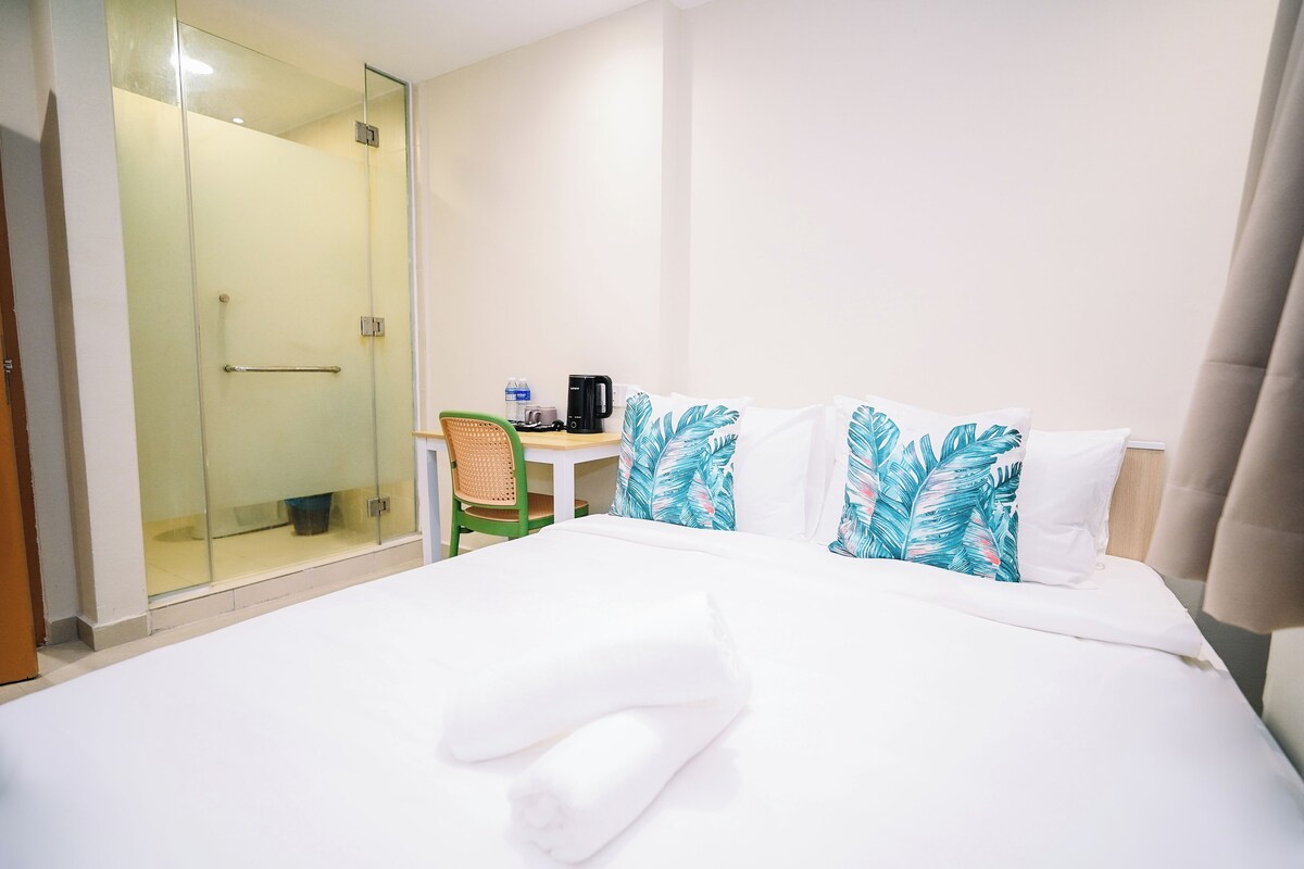 Habitación de hotel con cama, escritorio, silla, cafetera y cabina de ducha con paredes de vidrio.