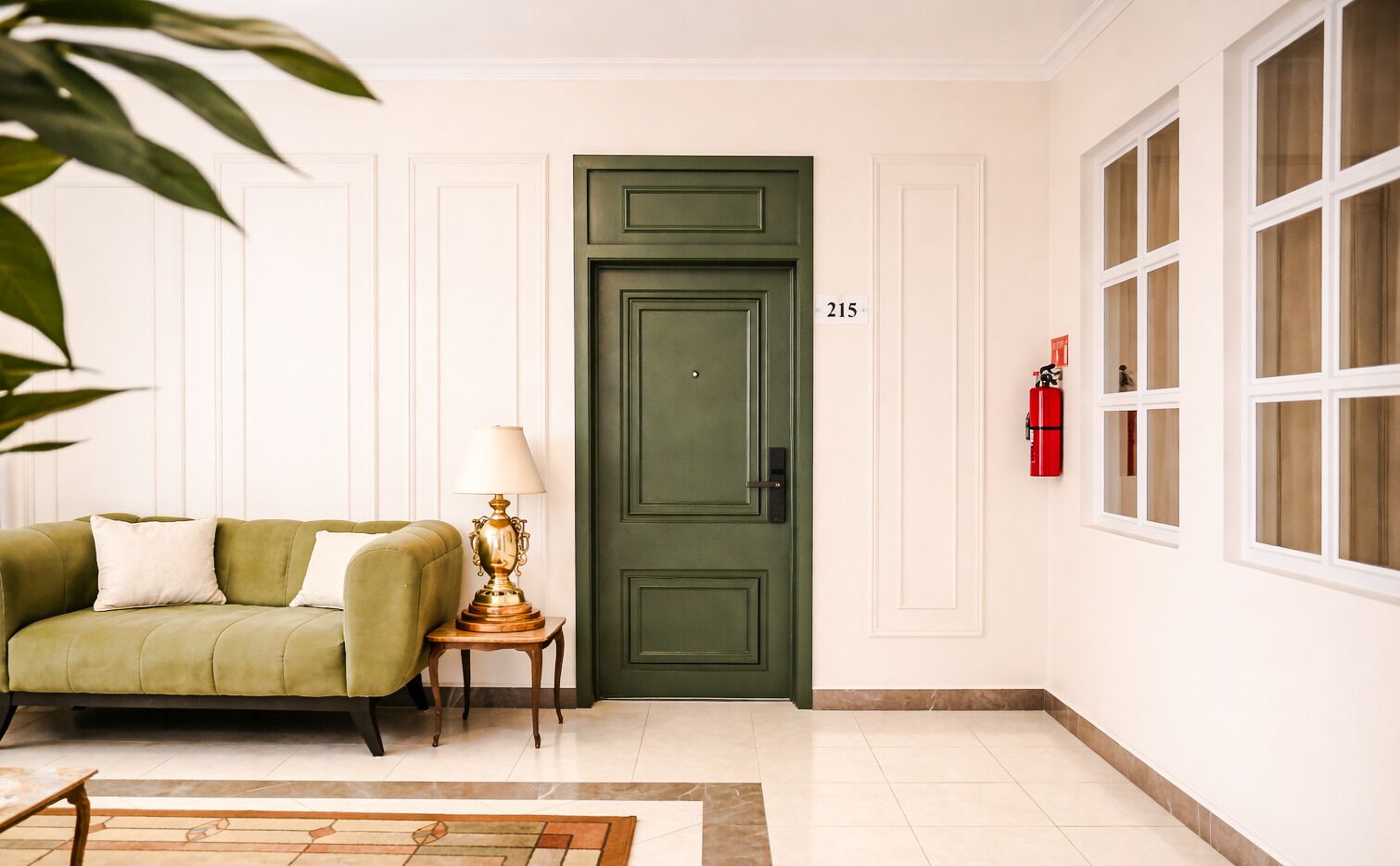 A room with a green door, a sofa, a lamp, a fire extinguisher, and a window.