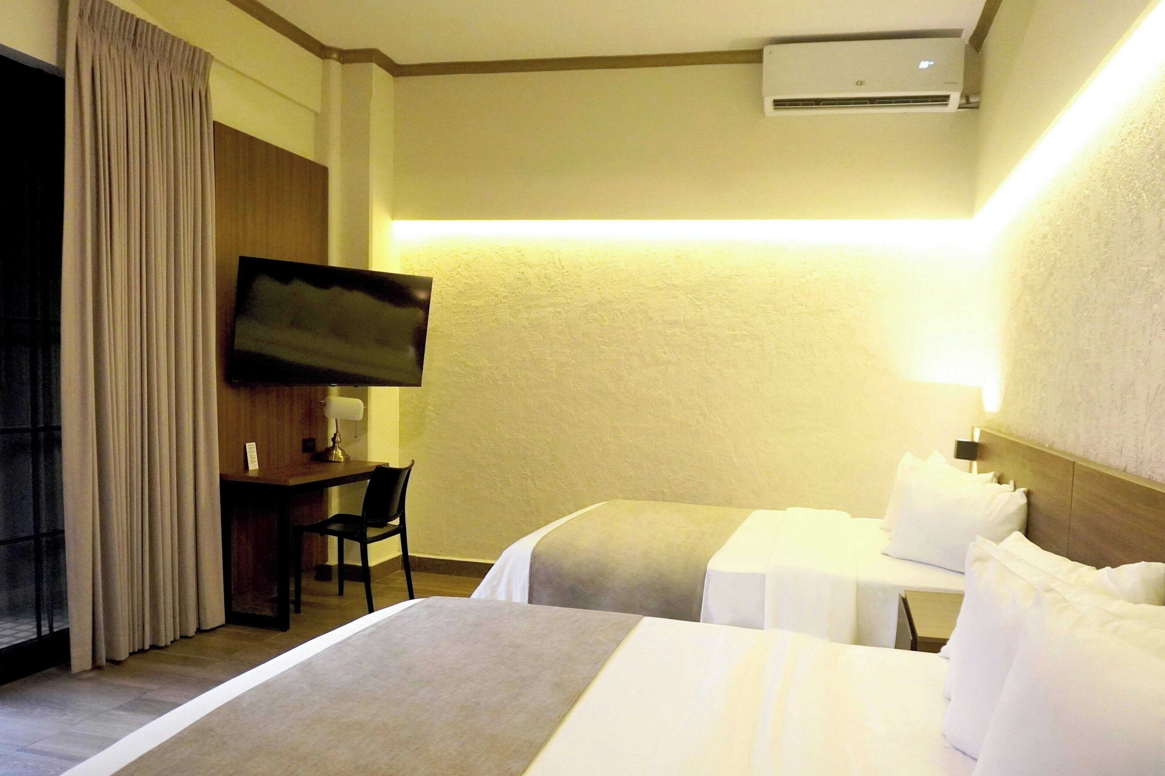A hotel room with two beds, a desk, a chair, a television, and an air conditioning unit.