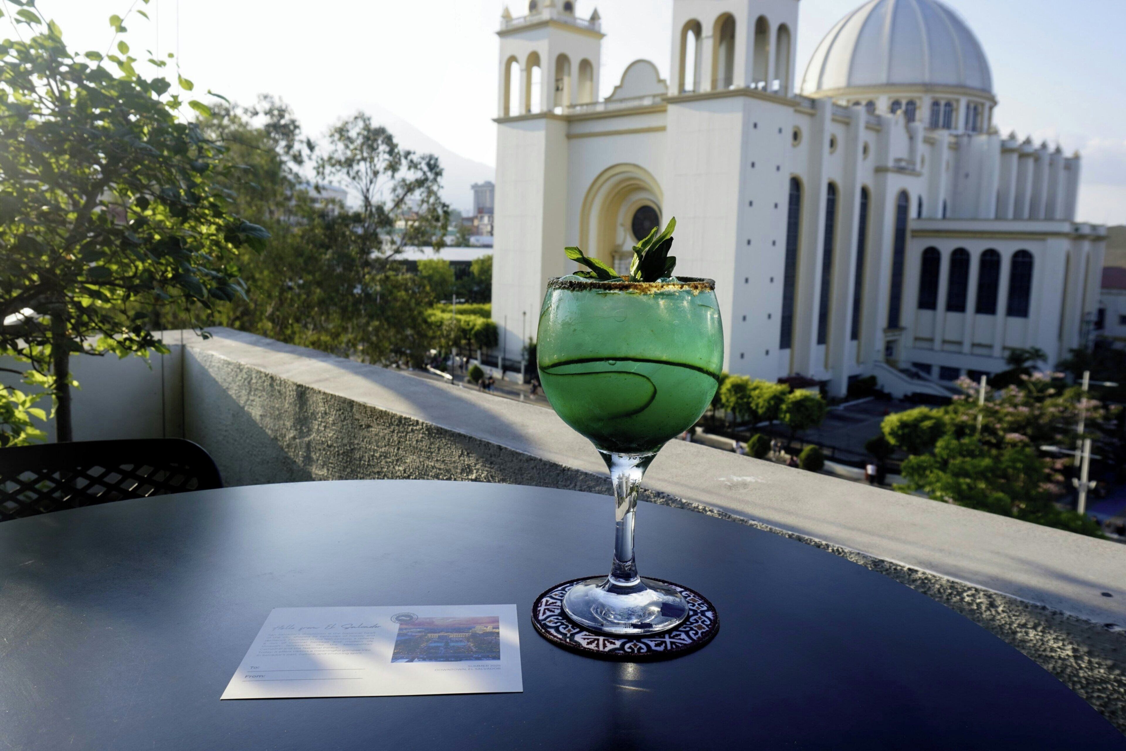 A cocktail glass with a green drink on a table with a postcard and a coaster.