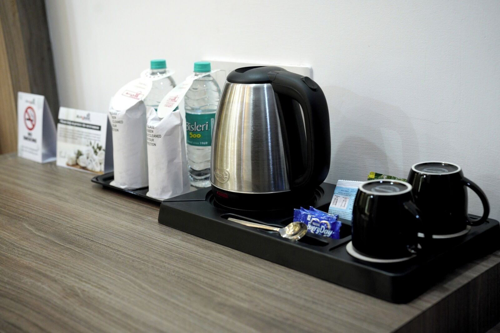 A hotel reception area with a coffee maker, two black mugs, a spoon, a packet of sugar, and two bottles of water on a tray.