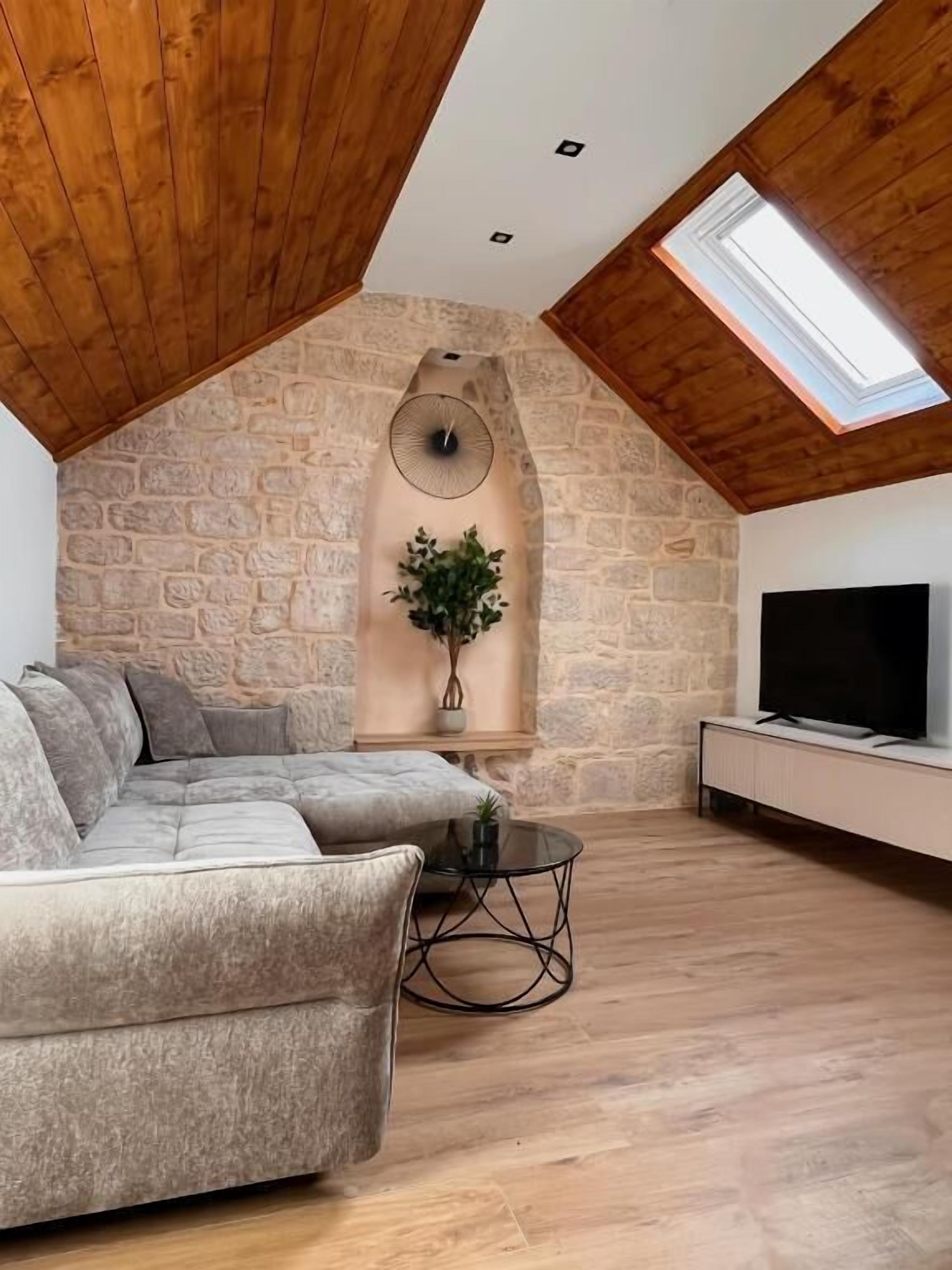 A loft-style room with a stone wall, a large window, a sectional sofa, a TV, and a small table.