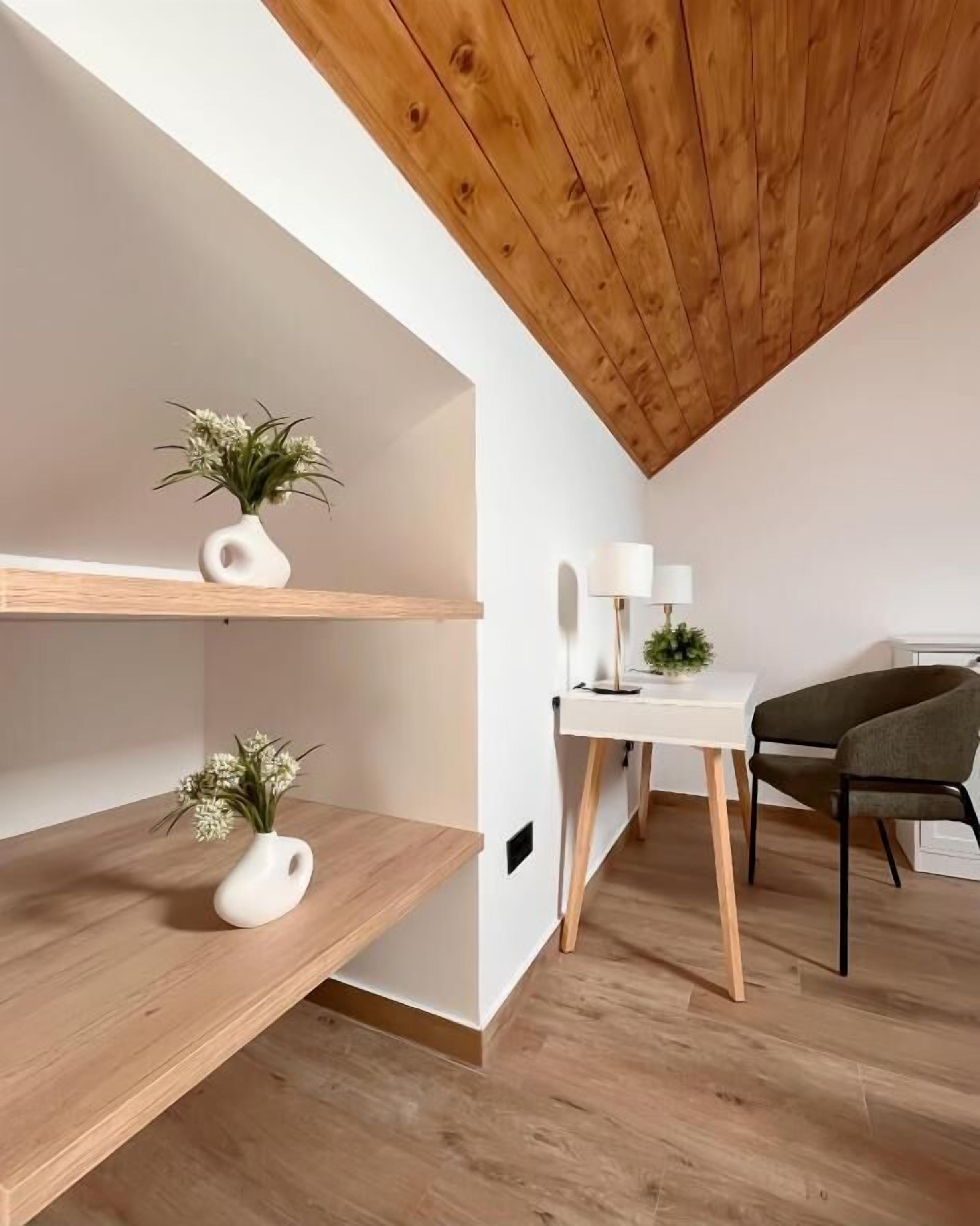 A modern loft bedroom with wooden beams, a desk with a lamp, and two white shelves with plants.