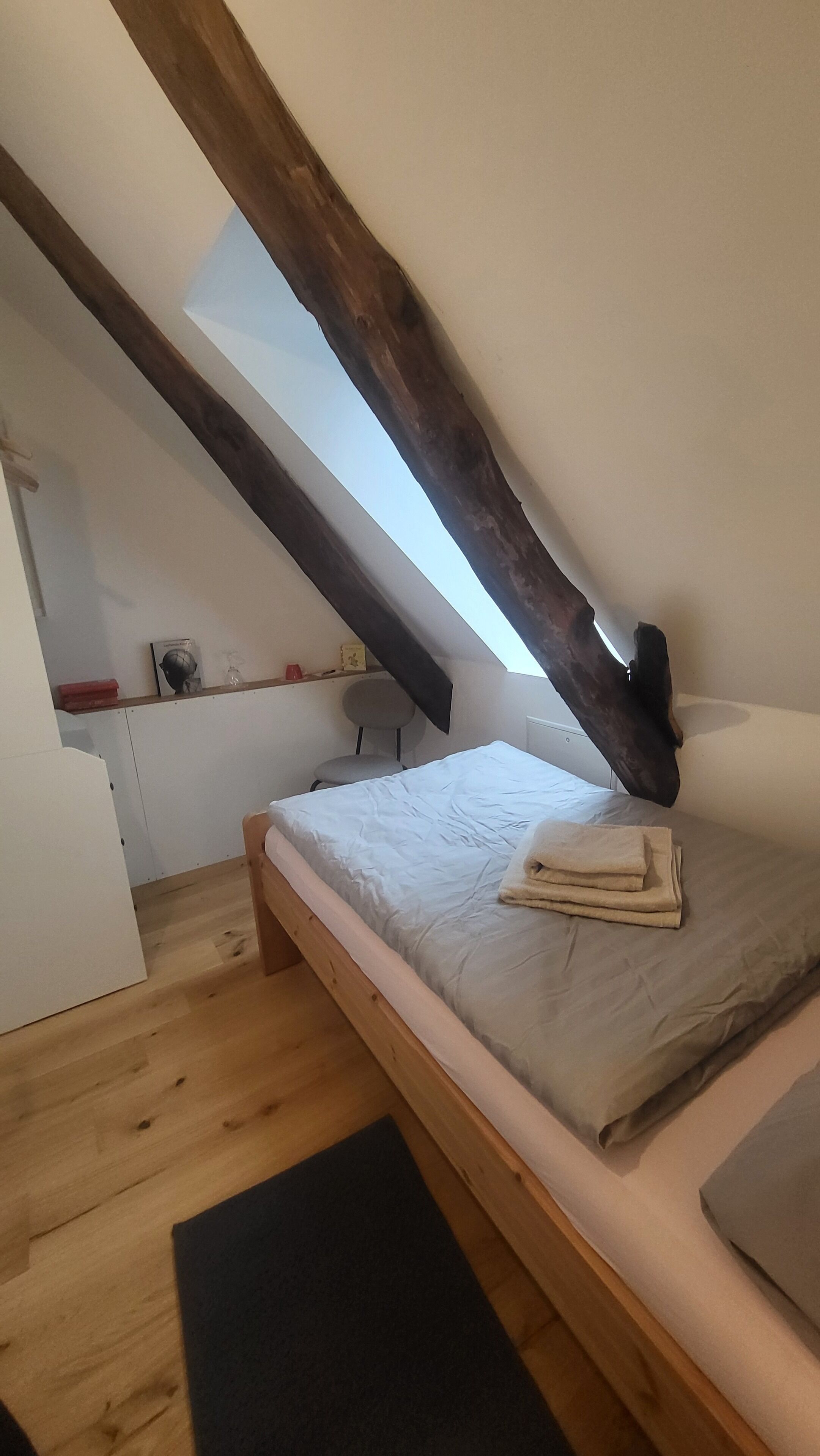 Une petite chambre mansardée, chaleureuse et accueillante, avec un plafond en pente, un lit simple, une chaise et une petite table avec une lampe.