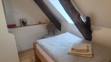 Une petite chambre mansardée, chaleureuse et accueillante, avec un plafond en pente, un lit simple, une chaise et une petite table avec une lampe.