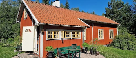 A red wooden house with a tiled roof, a bench, and potted plants in the yard.