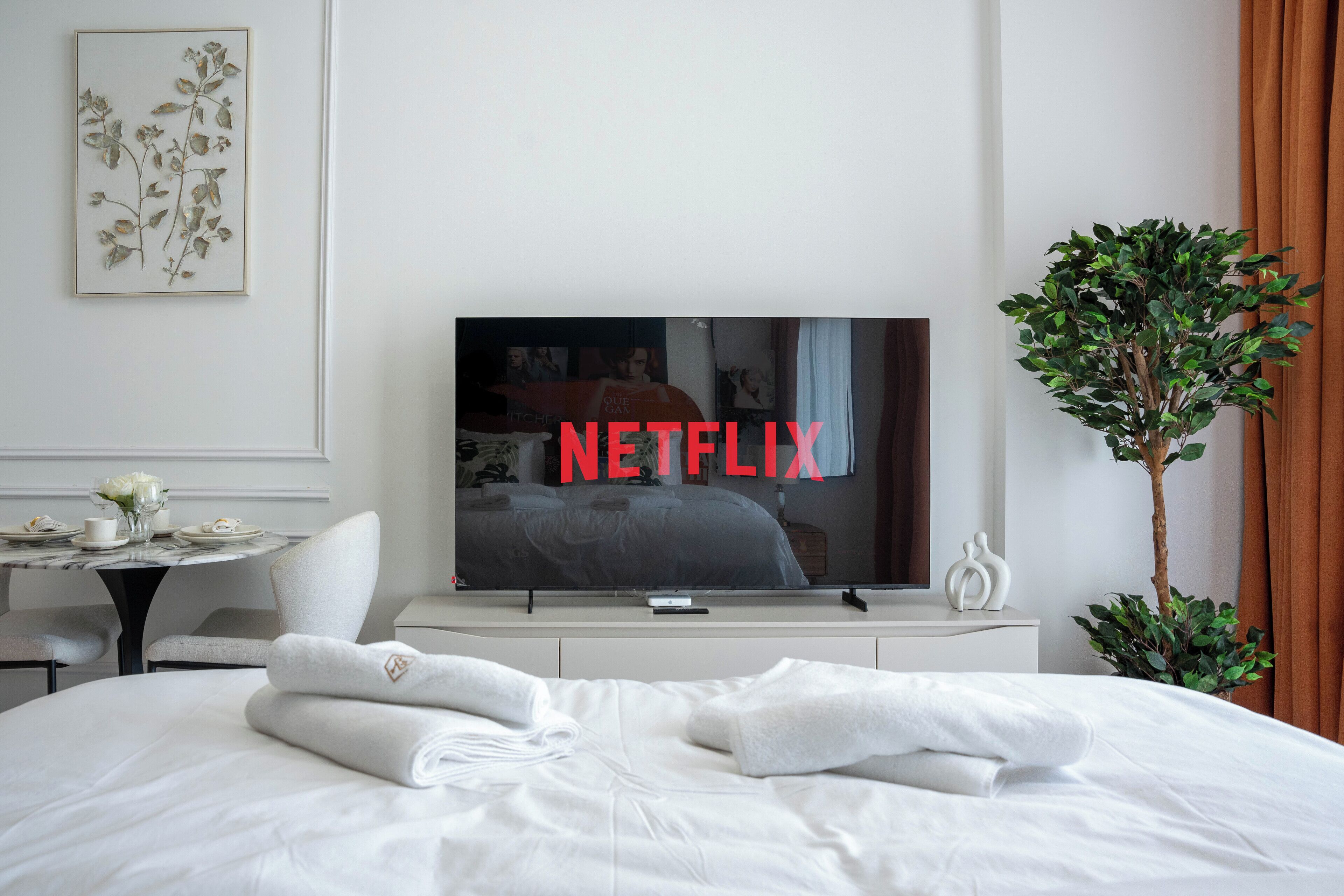 Une chambre d’hôtel avec un lit, un téléviseur affichant le logo de Netflix, une petite table avec un vase de fleurs et une plante en pot.
