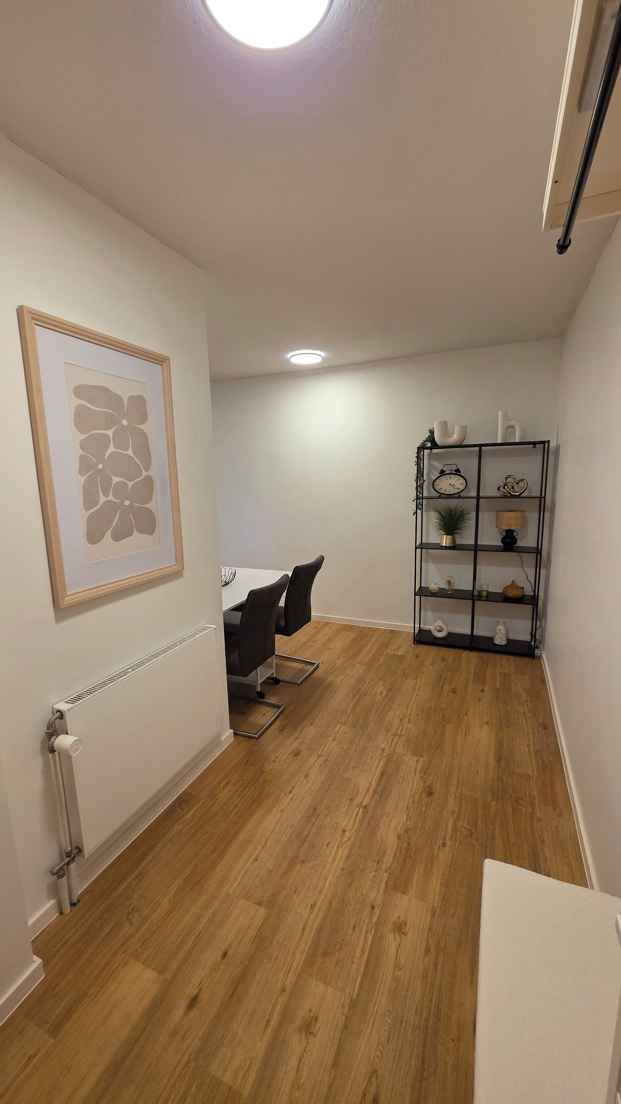 A room with a wooden floor, a white radiator, a framed artwork, a black chair, a white desk, and a black shelving unit with various items.
