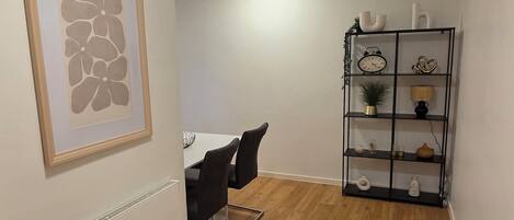 A room with a wooden floor, a white radiator, a framed artwork, a black chair, a white desk, and a black shelving unit with various items.