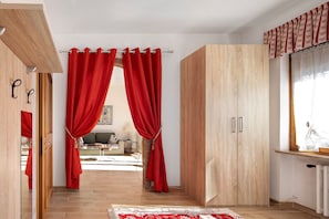 Ein Zimmer mit einem Holzschrank, roten Vorhängen, einem gemusterten Teppich und Blick in einen anderen Raum mit Sofa und Lampe.