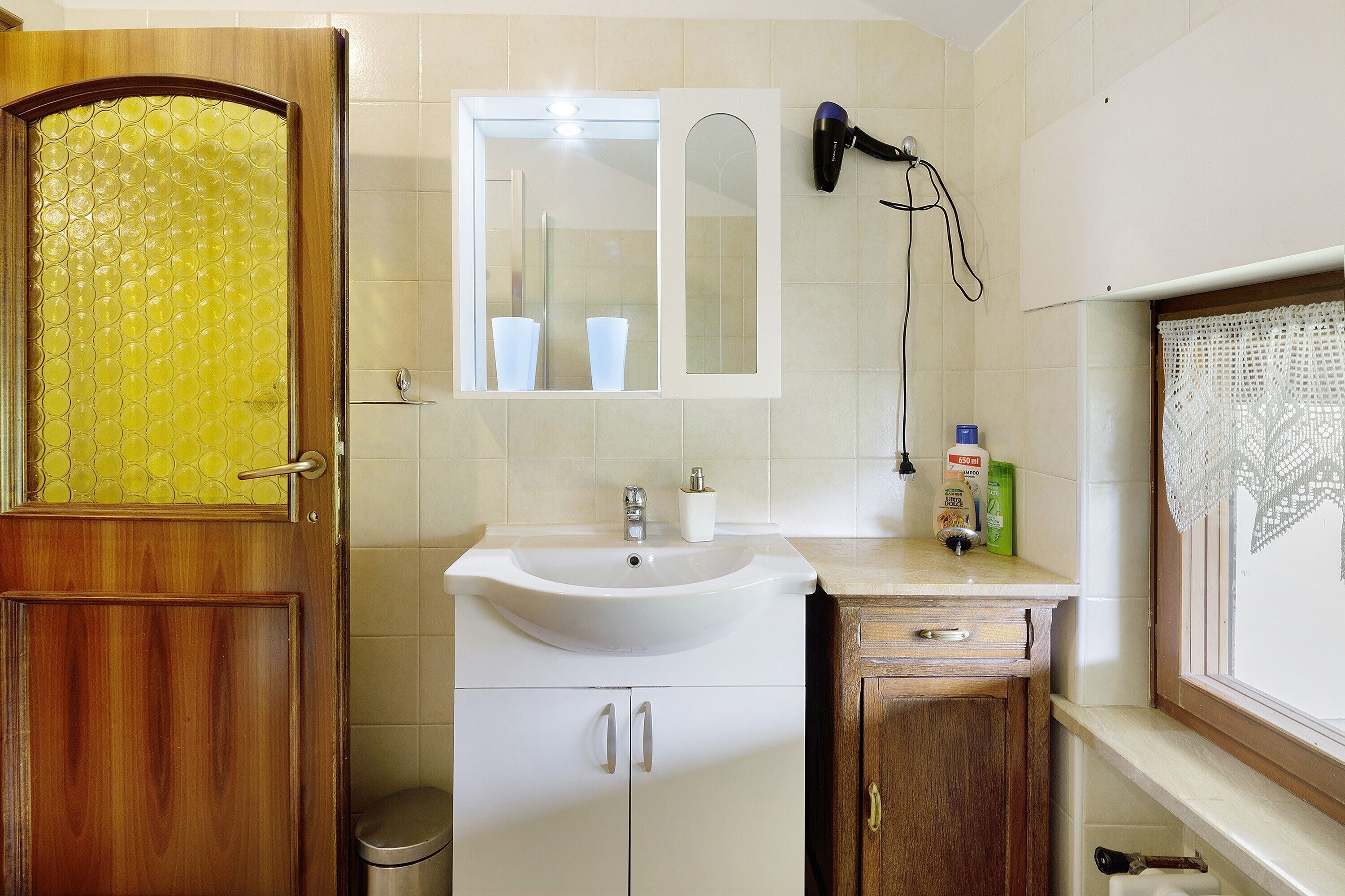 Ein Badezimmer mit Holztür, Waschbecken, Spiegel, Fön und einem Fenster mit Spitzenvorhängen.