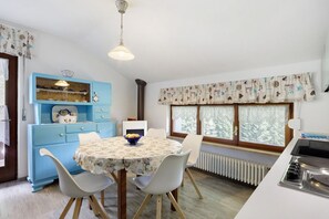 Un coin repas avec une table ronde, des chaises, un buffet bleu avec des étagères, une fenêtre avec des rideaux et un espace cuisine avec une cuisinière.
