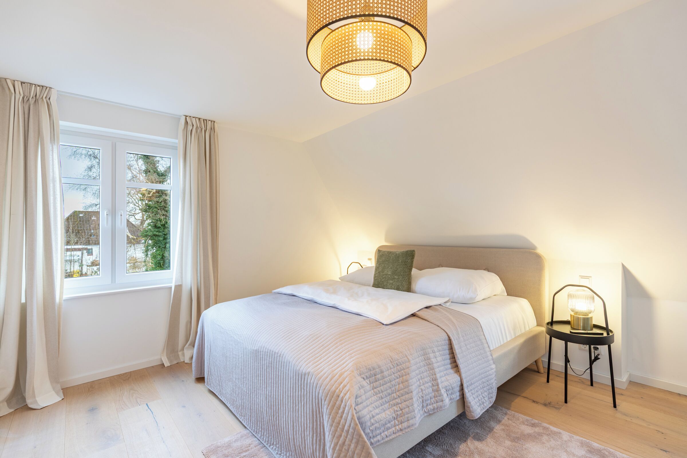 Ein Schlafzimmer mit einem Bett, einem Nachttisch mit Lampe, einem Fenster mit Vorhängen und einer Pendelleuchte.