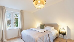 A bedroom with a bed, a bedside table with a lamp, a window with curtains, and a hanging light fixture. - Vacation home "Nordwärts" in Tönning - garden, fireplace & space for 6 guests (Tönning)