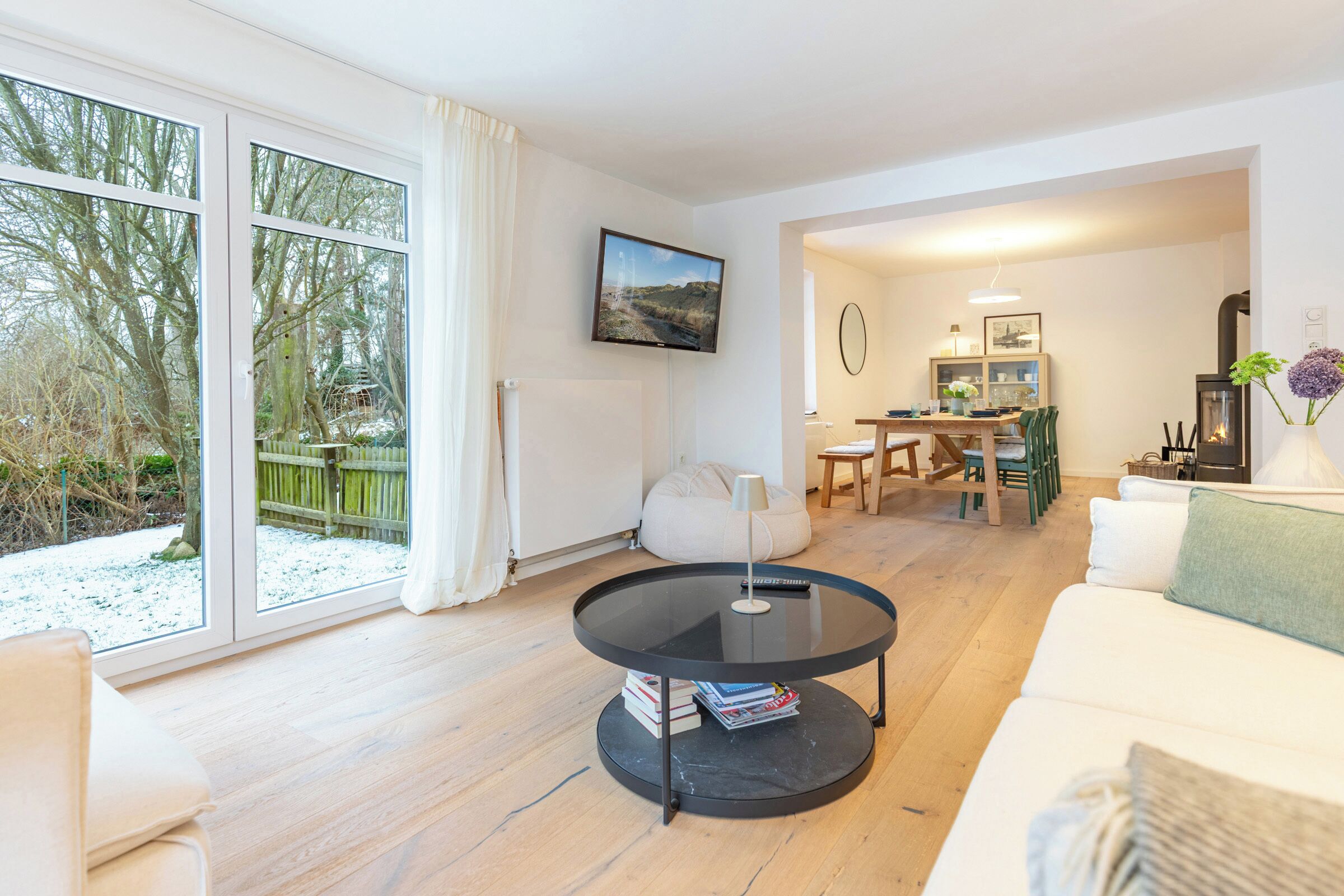 Ein modernes Wohnzimmer mit großem Fenster, einem runden schwarzen Tisch, einem weißen Sofa und einem Essbereich mit Holztisch und Stühlen.