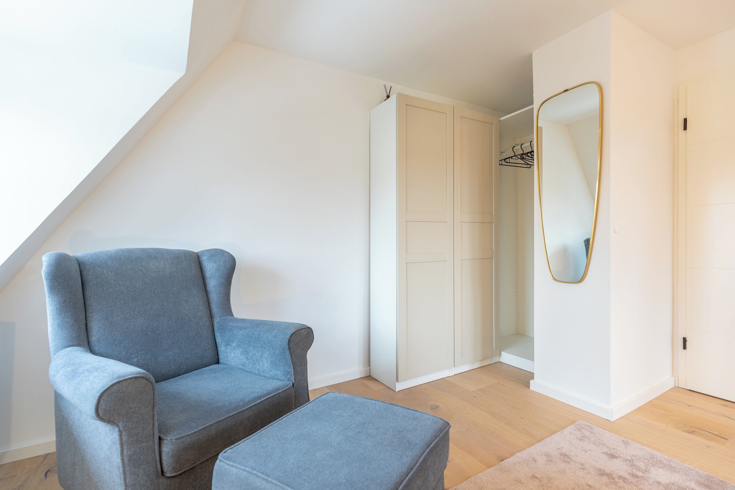 A room with a grey armchair and ottoman, a white wardrobe, and a large mirror with a gold frame.