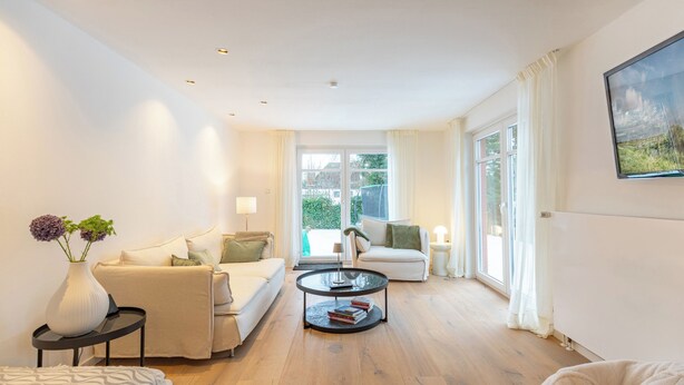 A modern living room with a sofa, a coffee table, and a lamp. There is a window with curtains and a painting on the wall. - Vacation home "Nordwärts" in Tönning - garden, fireplace & space for 6 guests (Tönning)