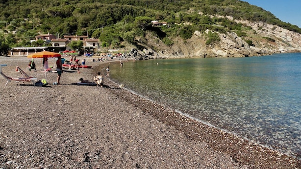 En strand med småsten, klart vatten, några parasoller och en del folk.