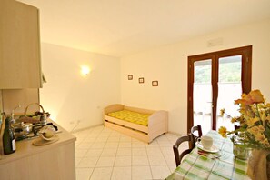 Ein kleines Zimmer mit einem Bett, einem Esstisch mit Blumen und einer Küchenzeile inklusive Herd, sowie einem Fenster mit Aussicht.