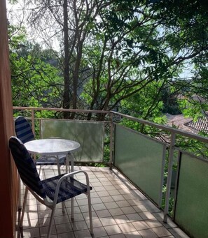 A balcony with a table and chairs, surrounded by a green mesh fence, overlooking a garden with trees and a tiled roof in the background. - Hotel Amica (Salsomaggiore Terme)