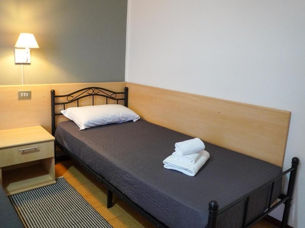 Una cama individual con cabecera de madera, una mesita de noche con lámpara y una toalla doblada sobre la cama.