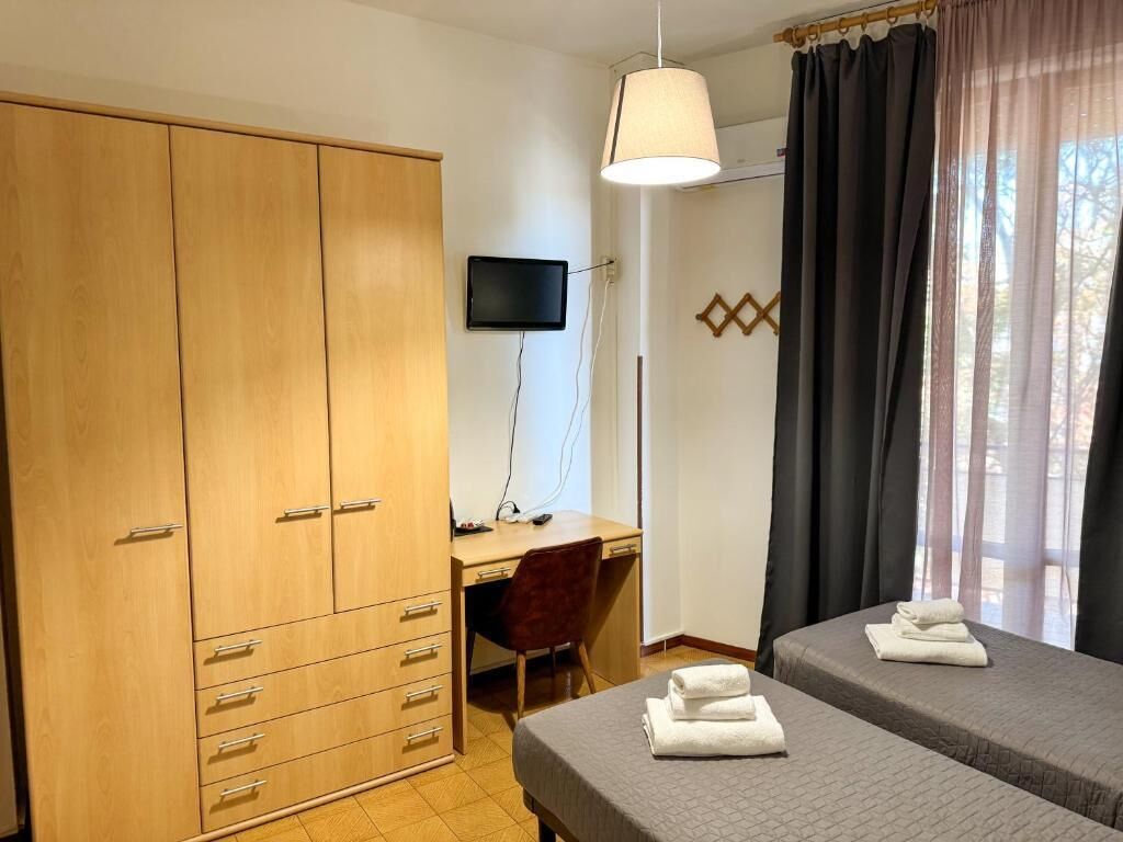 Ein Zimmer mit Holzschrank, Schreibtisch mit Stuhl, Bett mit gefaltetem Handtuch und Fernseher an der Wand.