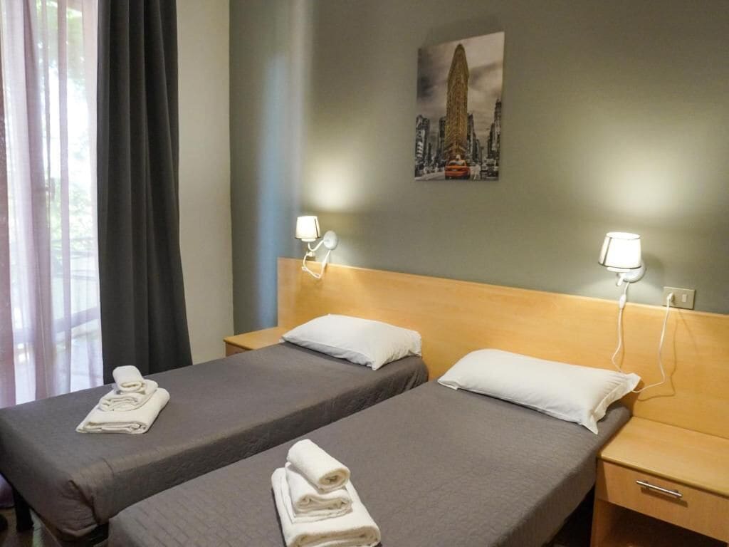 A hotel room with two single beds, each with a pillow and a folded towel. There is a nightstand with a lamp on each side of the beds. A painting of a cityscape is visible on the wall above the beds.