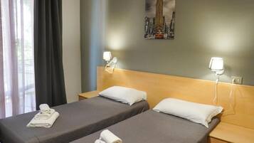 A hotel room with two single beds, each with a pillow and a folded towel. There is a nightstand with a lamp on each side of the beds. A painting of a cityscape is visible on the wall above the beds.