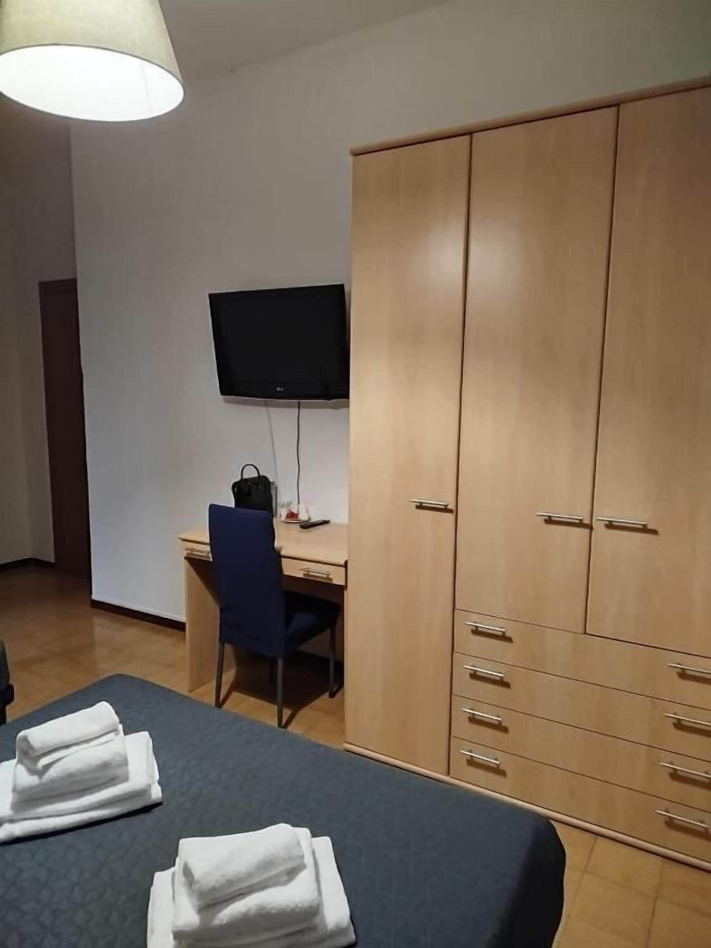 Quarto com cama, escrivaninha, cadeira, guarda-roupa e televisão na parede.