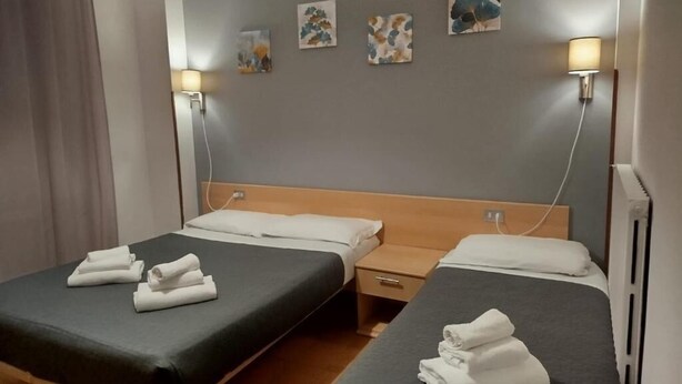 A hotel room with two beds, each with a neatly folded towel, a wooden headboard, bedside tables, and wall-mounted lamps. - Hotel Amica (Salsomaggiore Terme)