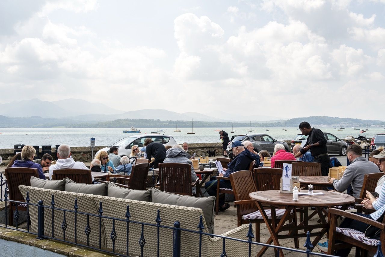 A waterfront café with outdoor seating, a view of the sea, and mountains in the background.