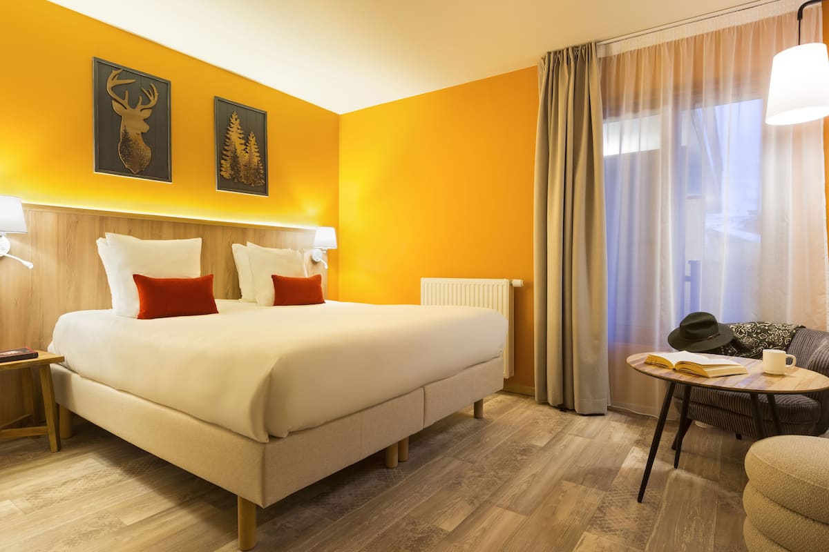 A hotel room with a large bed, a bedside table, a reading area with a chair and table, and a window with curtains.