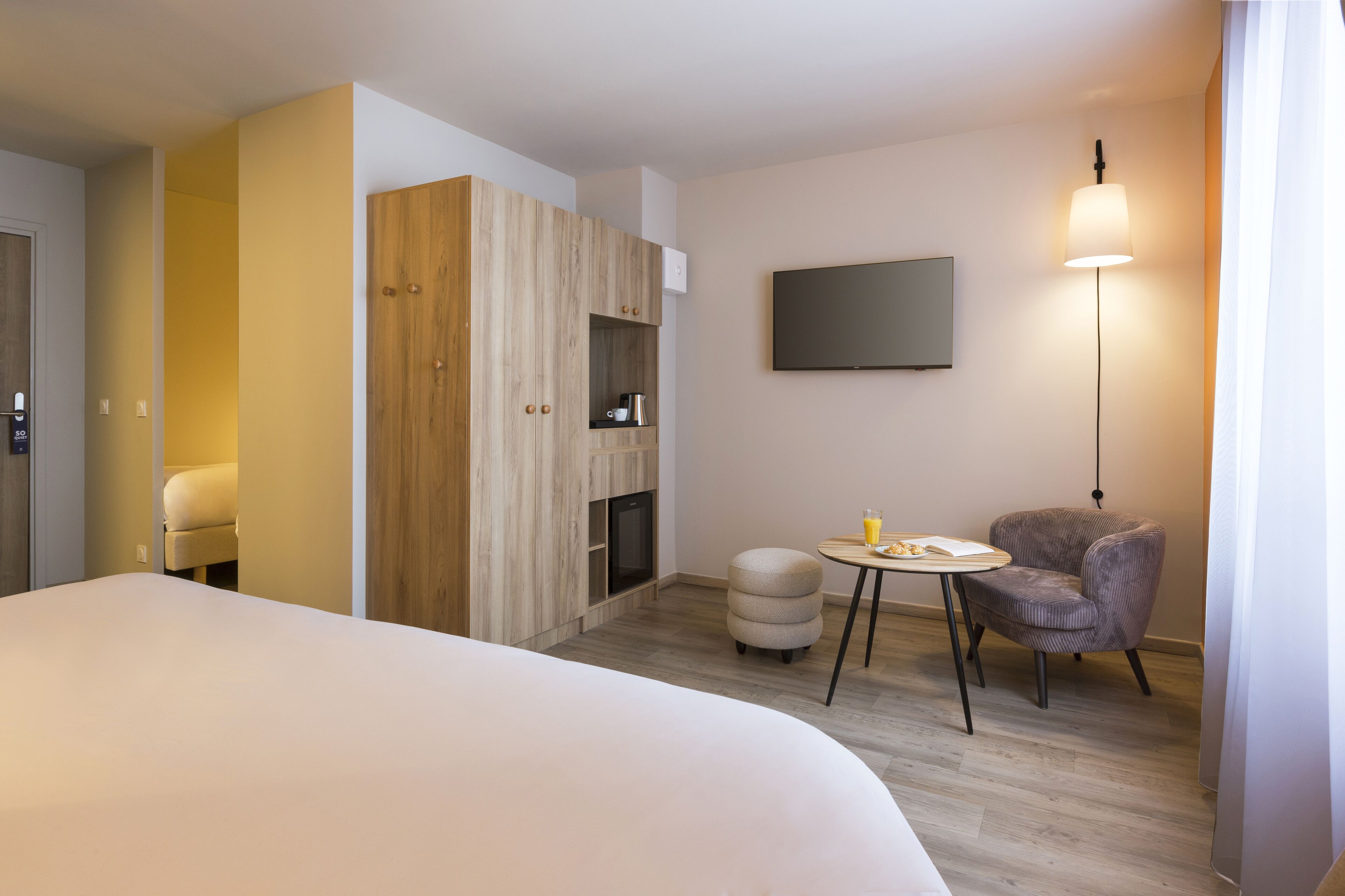 Une chambre d’hôtel moderne avec un grand lit, une armoire en bois, une petite table avec une chaise, un téléviseur à écran plat et un lampadaire.