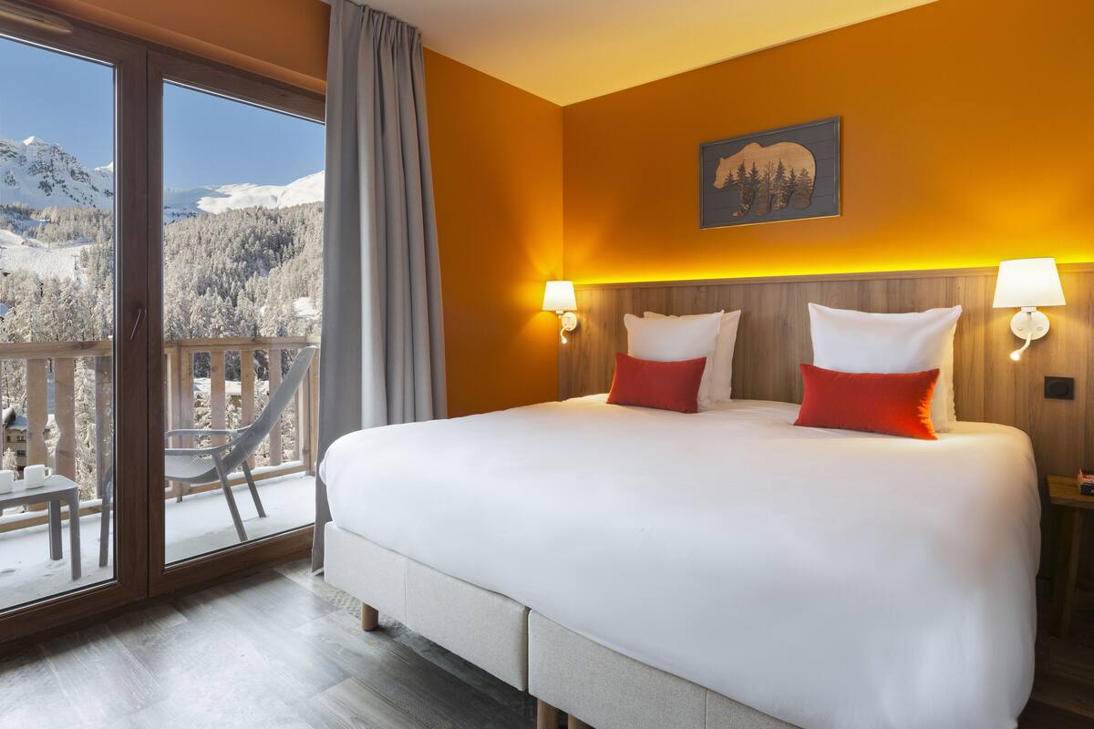A hotel room with a large bed, two bedside lamps, a wooden headboard, and a view of snow-covered mountains through a sliding glass door.